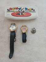 Two Looney Tunes Armitron wristwatches and a Tweety Bird locket on light fabric background, with original box showing Looney Tunes Armitron Collectibles branding.