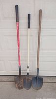 Photo of three shovels standing upright against garage door, showing different blade shapes and handle types.
