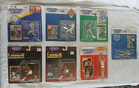 Photo showing all seven Starting Lineup packaged sports figure and card sets laid out on white surface. Includes baseball, basketball, hockey figures in various colored uniforms with cards.