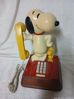 Photo showing front of vintage Snoopy and Woodstock telephone with Snoopy holding yellow handset and numeric keypad visible on red and brown base.