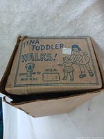 Photo of the top of the cardboard box showing the worn label with text 'Tina Toddler Walks' and the Imperial Crown Toy Corp logo, made in Brooklyn, NY. The box is visibly damaged with crumpled corners.