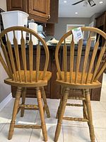 Rear view of two oak wood swivel barstools with rounded spindle backs and turned legs in a kitchen setting.