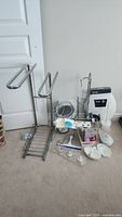 Various bathroom accessories including chrome towel stand, lit mirror, toilet paper holder with shelf, soap dishes, and makeup accessories arranged on floor