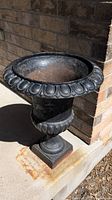 Full view of black cast iron urn planter showing its overall shape and weathered patina taken outdoors on concrete near brick wall.