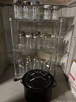 Metal shelving unit with three rows of clear glass mason jars in two sizes and a large black enamel canning pot on the floor in front.
