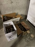 Box with various glass canning jars and metal lids in cardboard boxes shown on concrete floor.