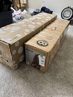 Two cardboard boxes for Christmas trees side by side on carpeted floor in a room with other household items.