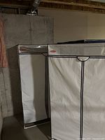 Two white Rubbermaid fabric zippered clothing storage racks against a basement wall, showing the front and side view of the units with metal frames visible underneath.