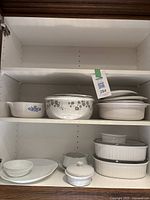 Shelf with multiple casserole dishes and bowls including Corningware with floral and blue cornflower designs along with some white ribbed dishes.