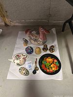 Overview of all decorative items arranged on a white cloth: large bird statue, small owl, teapot, salt and pepper shakers, two wooden painted plates, painted wooden egg, Ukrainian spoons.