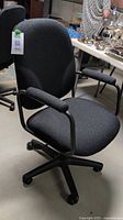 Front right angle view of black fabric desk chair showing cushioned seat, padded open armrests, and caster wheels.