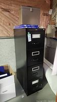 Full view of black 4-drawer metal filing cabinet with label holders, locking mechanism with key, placed in basement.