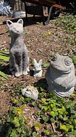 Four garden figures placed on the ground outdoors among plants including the concrete cat, frog, hobbit face, and resin gargoyle.