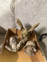 Overall view of the three garden stone figurines: two pigs and one gargoyle placed together in a cardboard box on concrete floor.