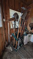 Wide view of wheeled garden tool storage stand holding multiple long handled garden tools including shovels, rake, broom, and hoe in a shed.