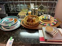 Photo of fruit themed bowls, wicker hot plates, wooden salt and pepper shakers, napkins and holder, wooden tray on kitchen counter.