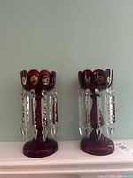 Front view of two cranberry glass luster candle holders each with 10 clear crystal spears hanging from scalloped rim, placed on white shelf against green wall.