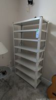 Full view of the white metal shelving unit with 8 shelves, placed against a wall in a room with carpet flooring.