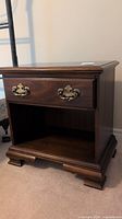 Full view of the wooden nightstand showing the single drawer with brass antique-style drawer pulls and open storage shelf below, sitting on bracket feet.