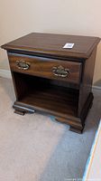 Full view of the nightstand showing single drawer with brass antique pulls and open shelf below, wooden finish with minor scratches.