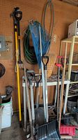 Photo shows assorted garden tools stored upright including shovels, rakes, broom, and pruner alongside green garden hose and shelving.