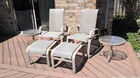 Wide front view of two chairs with footstools and side tables on a deck against brick wall.