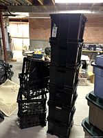 Stack of six black plastic storage crates with lids, standing upright in a basement environment, showing some dust and wear.