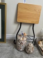 Fold-up wooden table resting against wall with two glass containers filled with seashells placed in front. Tiny blue glass whale figurine visible on the large container lid.