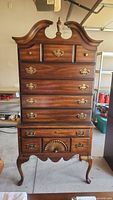 Front view of tallboy dresser showing broken pediment with finial on top, seven drawers with brass hardware, scalloped skirt, and Queen Anne legs.