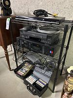 Wide view showing the Toshiba DVD and VHS players, JVC stereo cassette deck, power bar, and cassette tapes on black wire shelving.