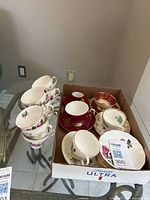 Set of various teacups and saucers placed in and beside a box, showing different floral designs and the Noritake red espresso cup.