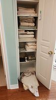 View of open linen closet shelves showing folded bath towels, mats, sheets, and linens, plus fabric tablecloths on floor.