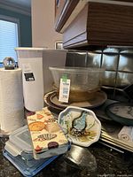 Photo of assorted kitchen items on a counter including paper towel holder with roll, white plastic wastebasket, glass mixing bowl with lid, decorative glass cutting boards with prints, tin box with seashell design, and kitchen clock.