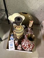 Overview of assorted vases including large white wavy rim vase, greenish-brown round vase, tall classical motif vase, reddish-brown patterned vases with bronze necks, and two small porcelain vases with floral patterns in a cardboard box.