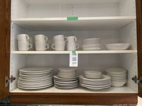 Shelf with stacked white dishes including mugs, plates, bowls, and saucers with ribbed cups visible.