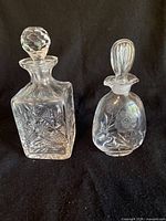 Two cut glass decanters displayed side-by-side on a black fabric background, showcasing their shape and design details.