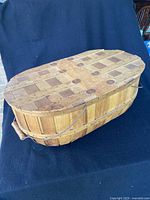 Closed view of the large oval picnic basket with hinged lid made of woven wooden slats and metal wire handle.