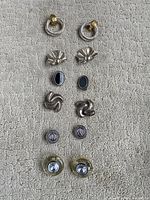 Full set of 5 pairs of clip-on earrings displayed on textured fabric background, showing different shapes and designs.