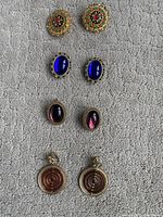 Top view showing four pairs of clip-on earrings on grey carpet: two pairs of oval cabochon earrings in blue and purple, one pair large round beaded earrings, and one pair circular spiral patterned earrings.