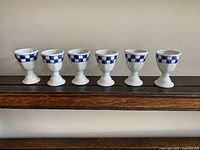 Front view of the set of six white ceramic egg cups with blue checkerboard pattern on upper rim, lined up in a row.
