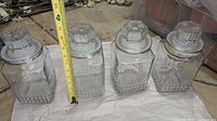 Four matching square glass jars with patterned sides and decorative lids measured at about 6.5 inches tall.