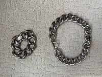 Photo showing the heavy silver-tone chain bracelet and necklace set laid out on a textured light gray fabric surface.