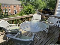 Full view of round dining table with four white resin wicker chairs and green cushions on patio deck, showing table's glass top and chair style.