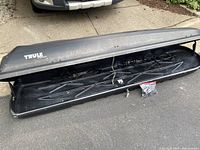 Wide view of the Thule Frontier rooftop cargo box in black with lid open, showing interior and included mounting hardware bag on pavement beside box.