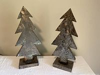 Front view of two handmade tin Christmas tree sculptures showing their textured metal finish and triangular layered design on square bases.