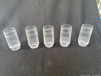 Front view of 5 transparent cut glass tumblers arranged in a row on black fabric background.