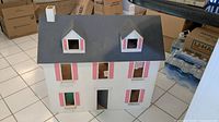 Photo showing front of vintage wooden doll house with dark gray roof, pink shutters, and open windows and door