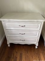 Front view of the cream-colored vintage dresser with three drawers and silver handles on wooden floor.