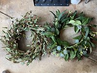 Top-down view of two artificial wreaths on a brown surface; one wreath features olive-like leaves, the other has green leaves with small white flowers.