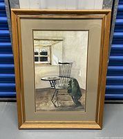 Framed print of a wooden chair with attached writing surface and green jacket near a window.